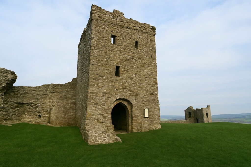 Stone tower of Llansteffan Castle with arched entrance and surrounding ruins on a grassy hill under a light blue sky.